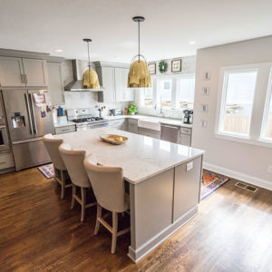 An all white kitchen with a large island with chairs and a single light hanging above it from the ceiling, windows all around, and stainless steel appliances