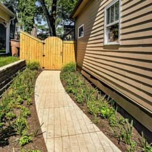 The side of a tan house with a newly placed path, surrounded by fresh landscaping, leading to a natural wood gate.