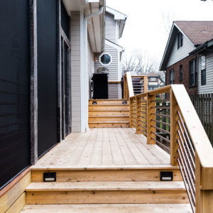 A natual wood porch on the side of the house with a narrow path leading to a grill.