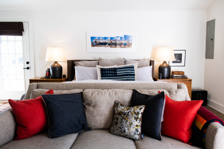 A master bed with an assortment of red and navy pillows at the end of it with two side tables with lamps and a picture centered above the bed