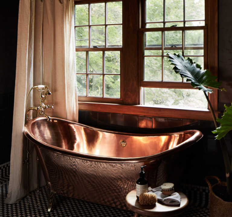 A tin tub in front of a wood-framed window with a curtain and plant beside it and a small table with lotion and a sponge