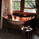 Gold tin tub in front of cracked open windows, green plant with a basket base, black and white checkered flooring, and cream curtain hanging behind