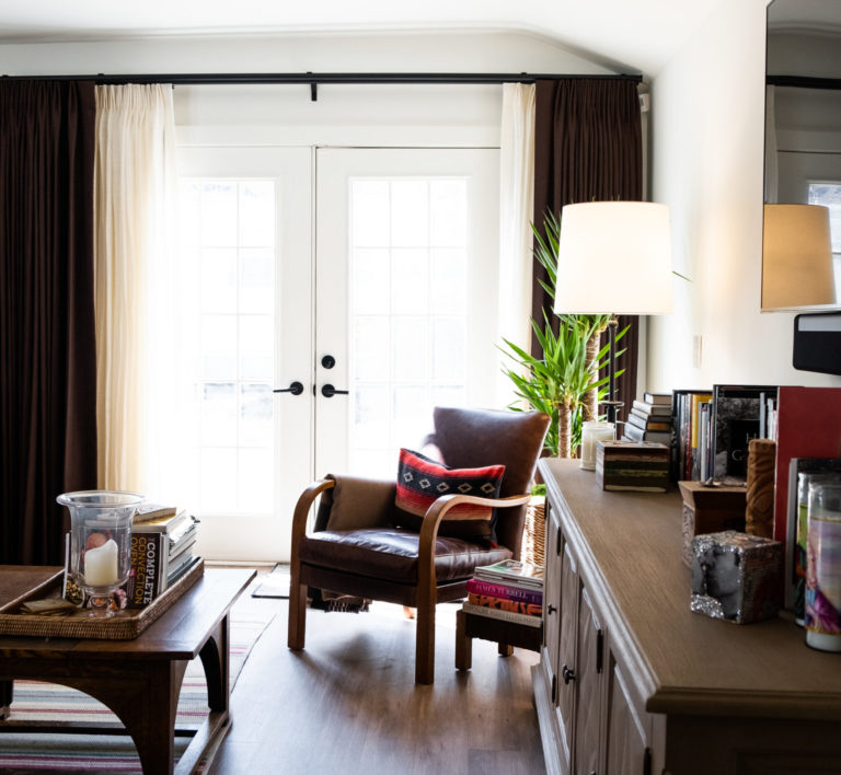 White room with french doors, brown curtains opened, dark brown leather chair with pillow, a dark wood dresser with books on top, small coffee table with a stack of books and a red, black, light brown rug underneath