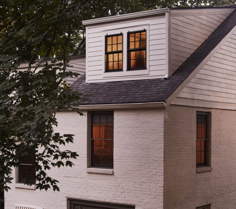 White house with black roofing, light shining through all windows, and green trees around