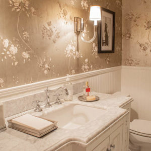 Cream colored bathroom with sink, toilet, lamp on the wall, and a picture frame on a wallpapered background