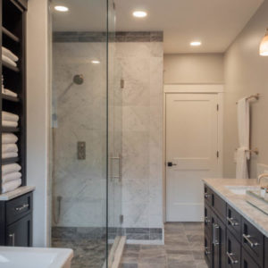 A bathroom with a large glass shower with grey tile trim around white tile, black cabinets with towels, a sink with black cabinets underneath, towel rack with white towels