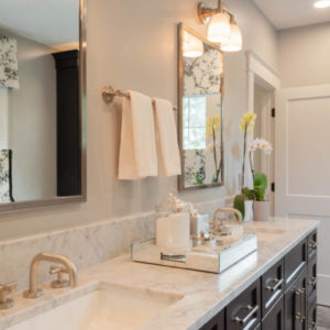 A bathroom with white granite counters, a double sink with mirrors above each, black cabinets below, a hand towel rack with white towels and an assortment of products on the center of the counter