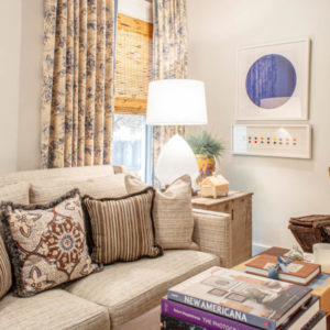 White room with cream couch and brown pillows, coffee table with a stack of books, floral curtain hanging on window behind the couch, a lamp turned on sitting on a nightstand with a picture of a purple circle in the background