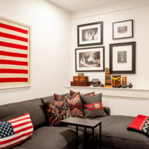 A grey sofa with an assortment of red and grey pillows, an American flag pillow, and the stripes of a flag picture on the wall above it