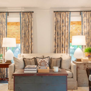 Cream room with a cream sofa and pillows in front of two windows with flowered curtains pulled back, side tables with lamps on both sides, and a coffee table with a stack of books in front of the sofa