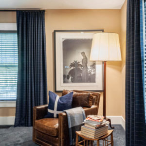 A room with blue curtains and a black and white picture on the wall with a brown leather chair and small sidetable with a stack of books in front of it