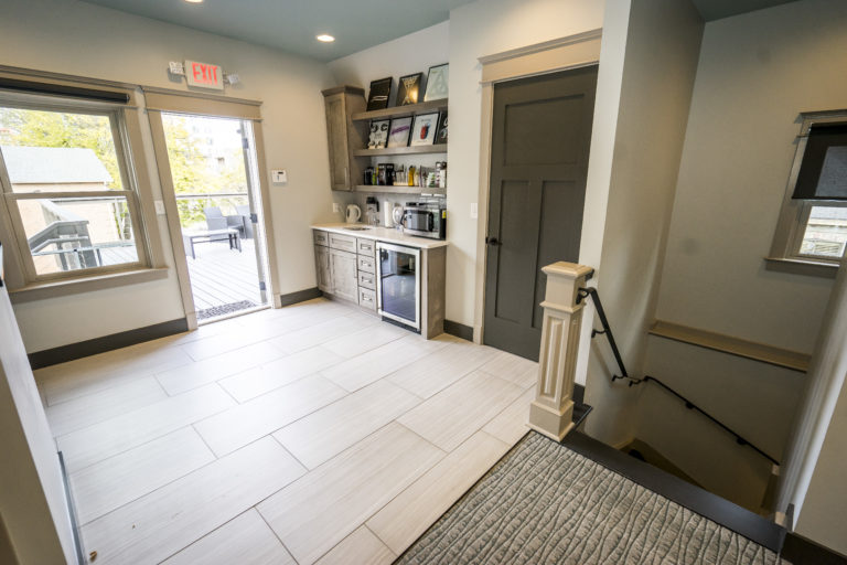 Room with bar and mini fridge, three shelves above with pictures, black closet door, door to patio with exit sign above, light flooring, staircase going down with grey carpet