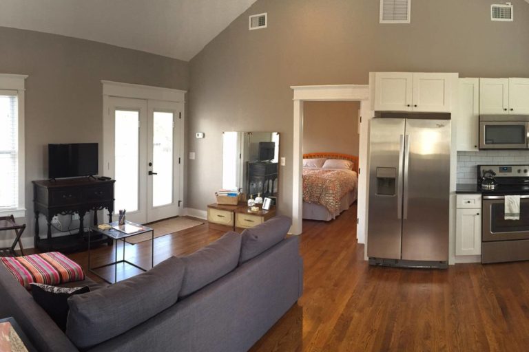 A living room with a grey sofa, black coffee table, french doors, and a stainless steel refridgerator with a master bedroom with orange bedding through an open door