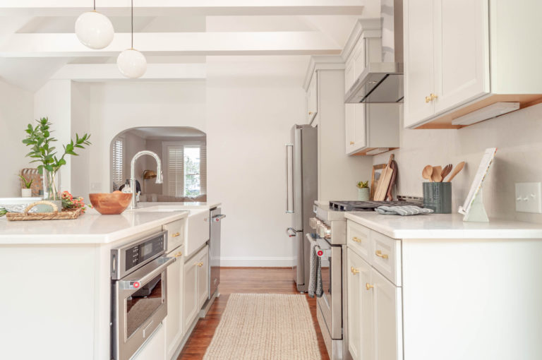 An all white kitchen with stainless steel appliances, round lights hanging from the celing of the island and a cream rug on the wood floors