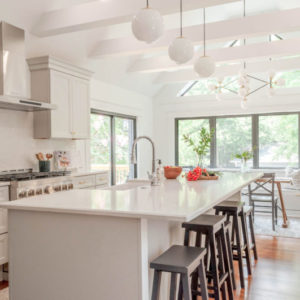 All white kitchen with three round bulb lights above an island with dark wood stools, white beams in the ceiling, five large windows, and light wood floors