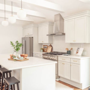 White kitchen with stainless steal oven, vent, and refridgerator, white island with grey stools, and green plant as the center piece