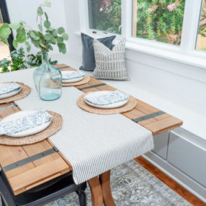 Wood table that is set, windows with white cabinets across the bottom, and grey pillows