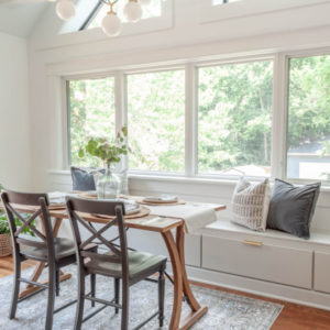 White room with three windows side by side looking at green trees, window seat in front of window with drawers underneath, brown table in front with grey chairs and plant on the center of the table