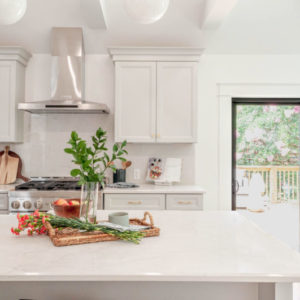 An all white kitchen with a large island counter with a plant on a wood cutting board, a glass door to the right and a stove and cabinets to the right