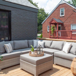 A grey L-shaped sofa on a deck with a coffee table and red brick house behind it