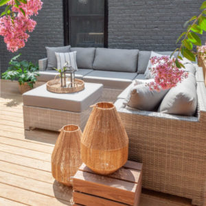 Deck outside grey brick house with light brown l-shaped wicker furniture and grey cushions, small coffee table, pink flowers around the sides