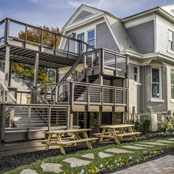 Grey home with two levels and decks and two picnic tables out front with flowers on the ground