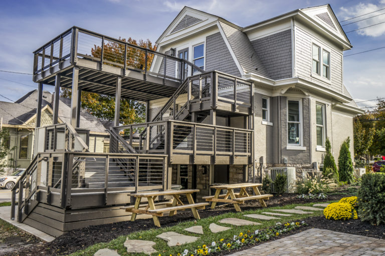 Grey home with two levels and decks and two picnic tables out front with flowers on the ground