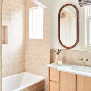 White bathroom with cream tile shower, oval mirror with wood trim, sink underneath a window with curtain rolled up, and light wood cabinets underneath