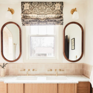 White bathroom with oval mirrors with wood trim to the left of a sink with a window with curtain rolled up, and light wood cabinets underneath