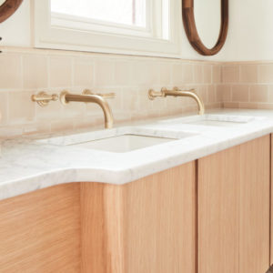 Double sink with light wood cabinets and gold faucets, white granite countertops, and cream backsplash