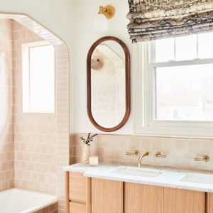 White bathroom with cream tile shower, oval mirror with wood trim, sink underneath a window with curtain rolled up, and light wood cabinets underneath