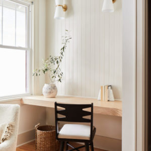 White room with desk and black chair with two lights above on the left and right, a plant on the desk and a basket underneath