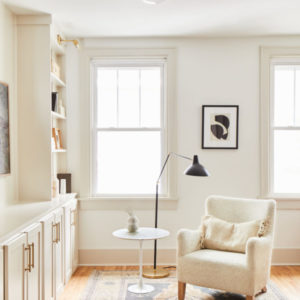 All white room with white book shelf and cabinets, chair, side table, two windows in the background, black lamp, and picture hanging on the wall