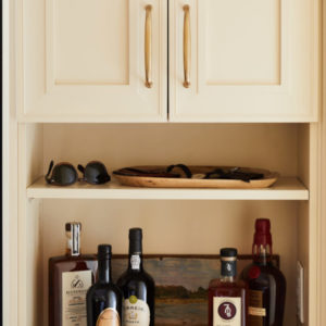 A cream-colored cabinet with sunglasses and liquer bottles on the open shelves