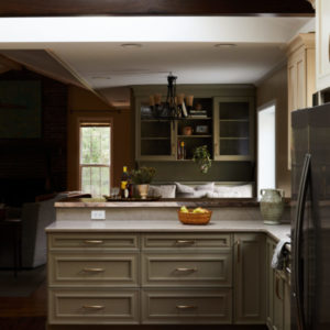 A dark picture of a kitchen overlooking cabinets into kitchen dining room with a chandelier and plate cabinet in the background, a stainless steel refridgerator to the right, and a grey chair in the sitting room to the left.