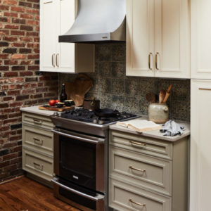 A brick wall with a stainless steel stove in front of it with white cabinets around, various kitchen utinsels lay on the counters