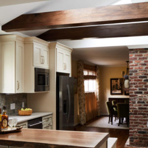 Kitchen with dark wood beams, white cabinets above a stainless steel refridgerator, grey backsplash, brick wall, looking into dinning room with table and chairs