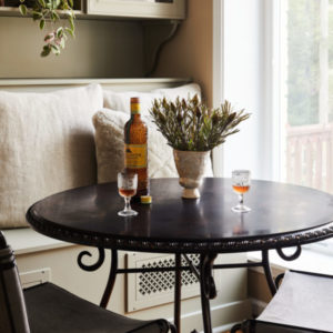 A black round table with two chairs, an open bottle and two drinks, green plant as a center piece, a light grey sofa behind it