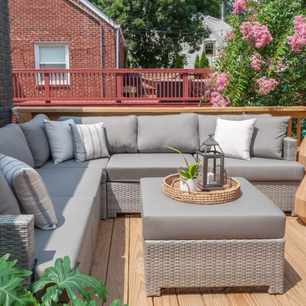 Patio with light gray couch and ottoman and a tree with pinks flowers in front of a red deck and brick house