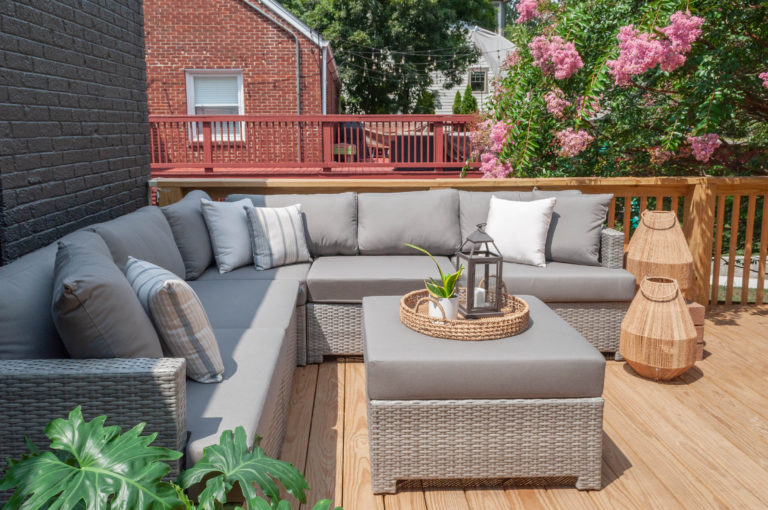 Patio with light gray couch and ottoman and a tree with pinks flowers in front of a red deck and brick house