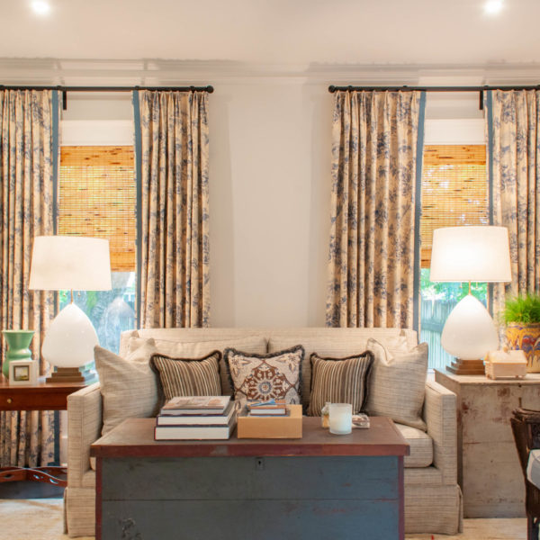 Cream couch with three pillows and a table on both sides with large white lamps in front of windows with floral curtains