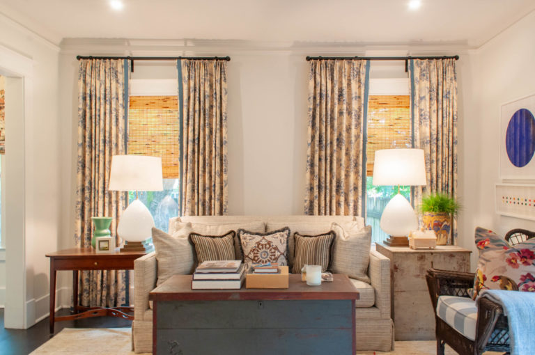 Cream couch with three pillows and a table on both sides with large white lamps in front of windows with floral curtains