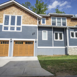 A blue and light wood home with wood door double garage and green trees behind it