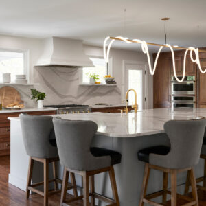 A full shot of a new kitchen remodel. The cabinets are white with a dark wood front and have a white marble countertop. The kitchen has a custom Luke Lamp overhead light.