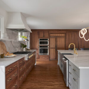 An open kitchen with custom wood and white cabinets with white marble counter tops. Across the room is custom wood cabinetry, with multiple pantries and double stacked oven.