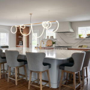 A white island remodel with a white marble top surrounded by gray fabric chairs and a custom-made overhead light. At the front of the island is a sink with a gold faucet.