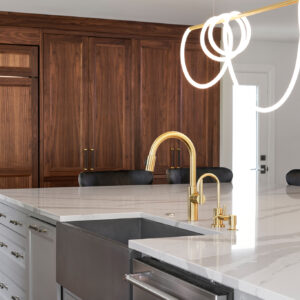 An upclose shot of the front of the custom island with a clear view of the attached sink with gold faucet and handles. In the background is the dark wood custom kitchen pantry.