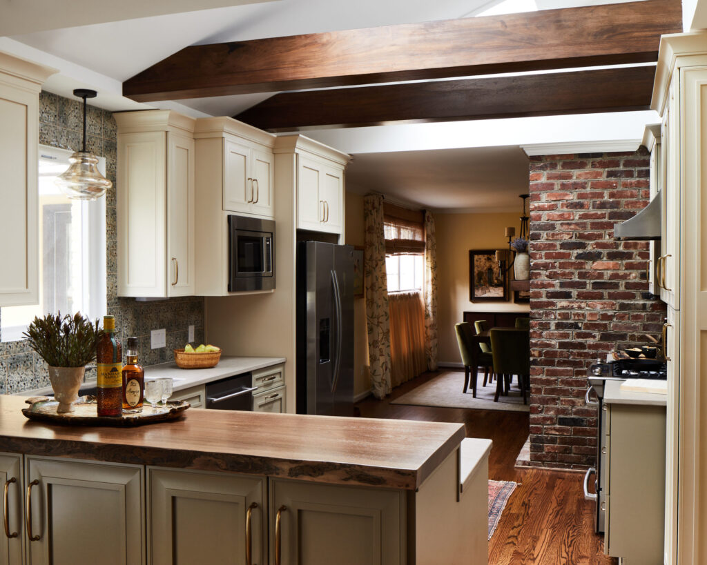 A modern kitchen featuring wooden ceiling beams, an island countertop, and a brick accent wall leading to a dining area.