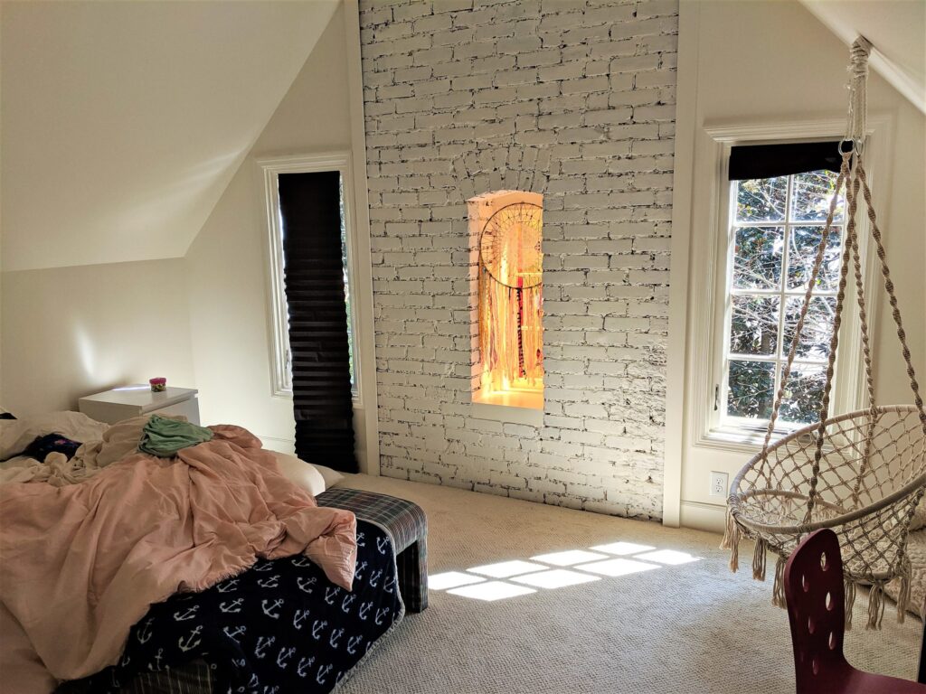 Cozy attic bedroom with a whitewashed brick wall, a hanging chair, and natural light streaming through a large window.