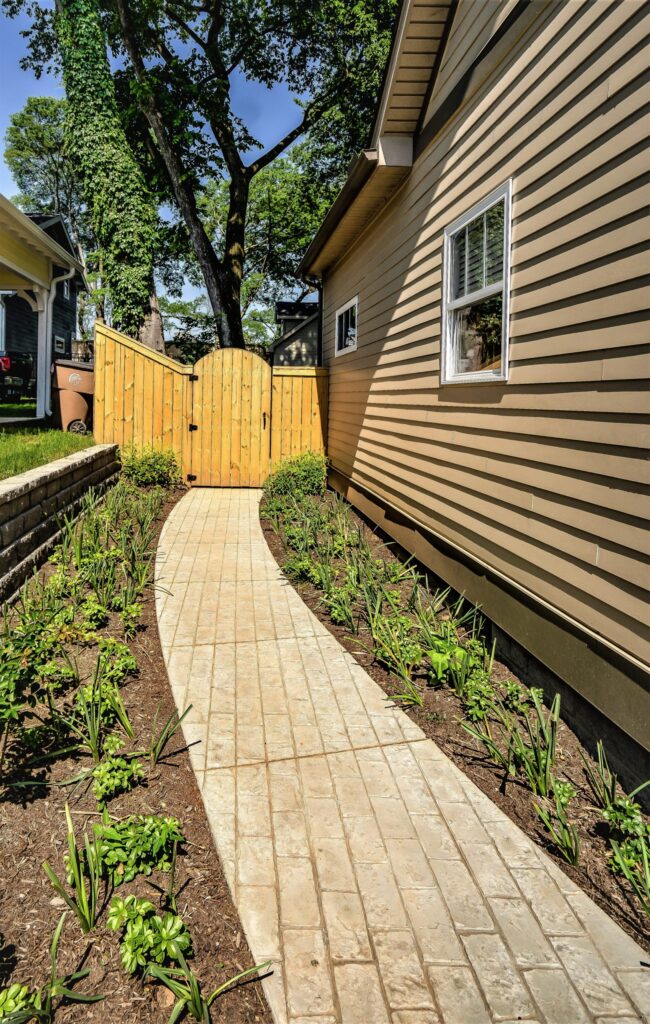 A paved pathway leads to a wooden backyard gate surrounded by plants.